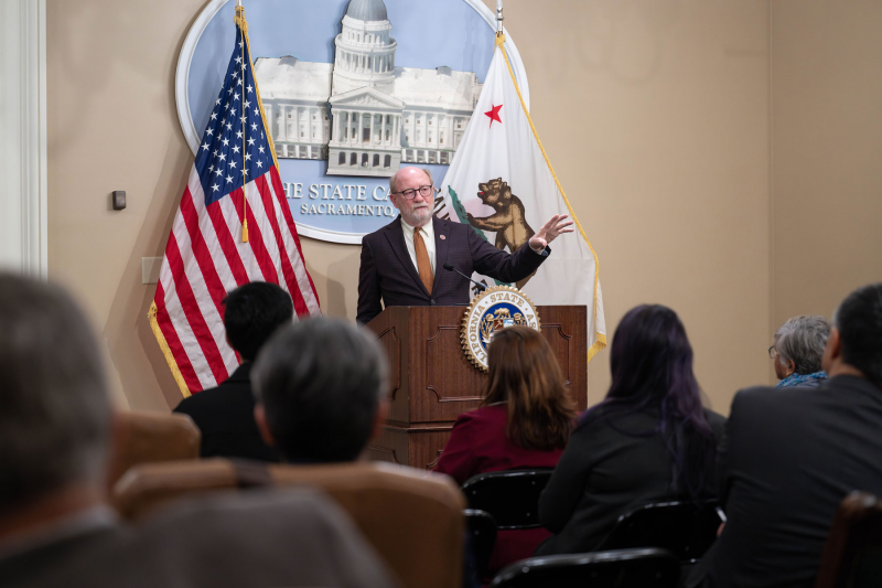 Senator Laird standing in a legislative room in Sacramento speaking at a podium to a crowd of local elected from Assembly District 29.)